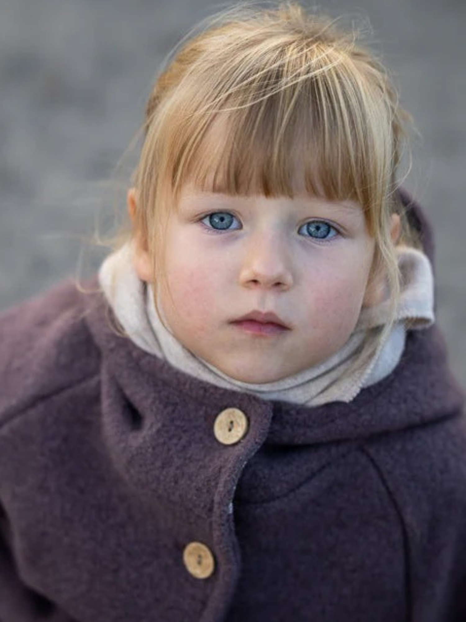 Das Bild zeigt ein Kind in einer Wichteljacke Wollwalk - Malve mit Holzknöpfen und Bio-Jersey-Futter, das direkt in die Kamera blickt, warm und bequem für Herbst bis Frühling.