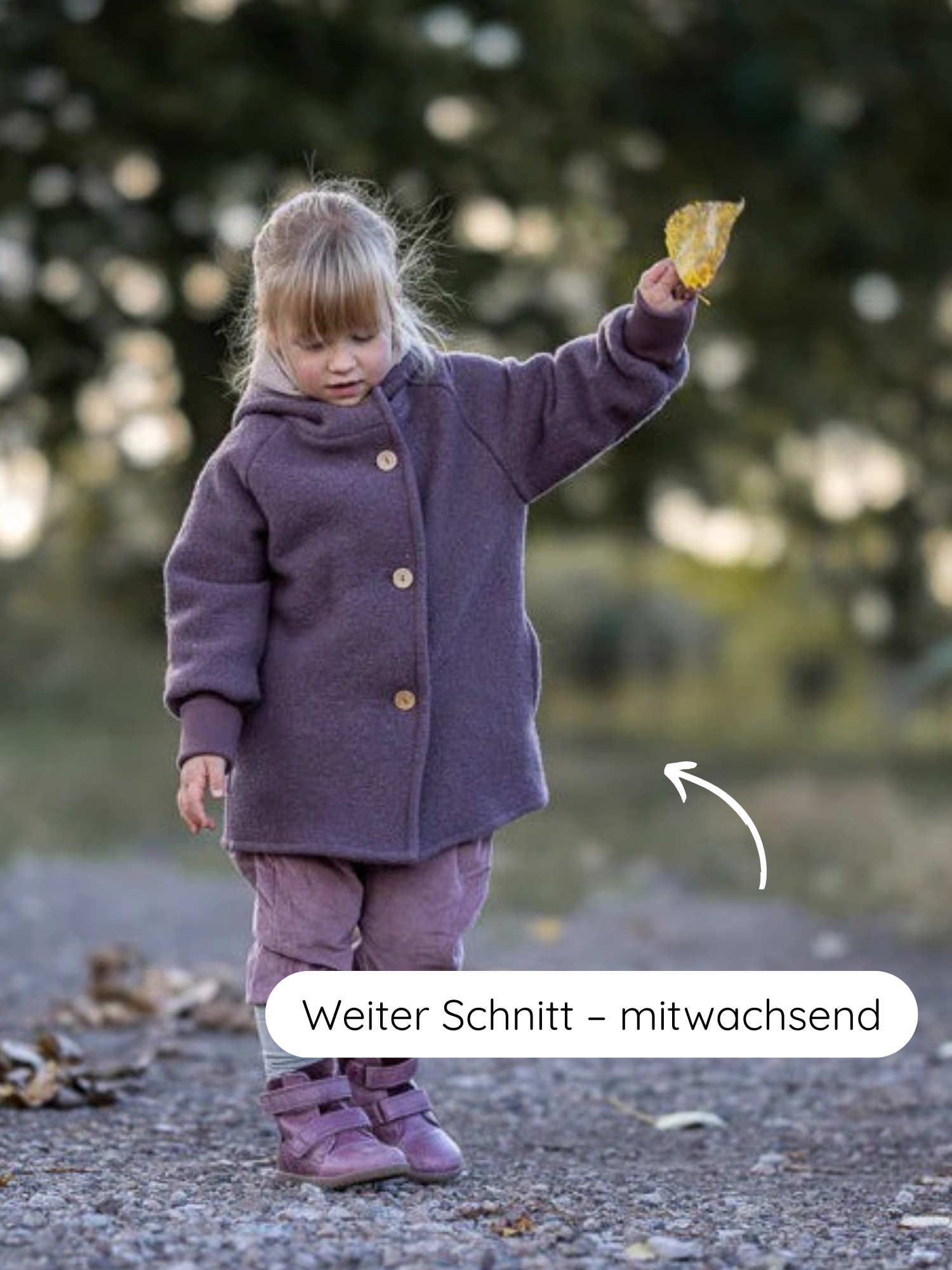 Ein Kleinkind steht auf einem Kiesweg und trägt die Wichteljacke Wollwalk - Malve mit großzügigem Schnitt, ideal für Herbst. Es hält ein gelbes Blatt hoch.