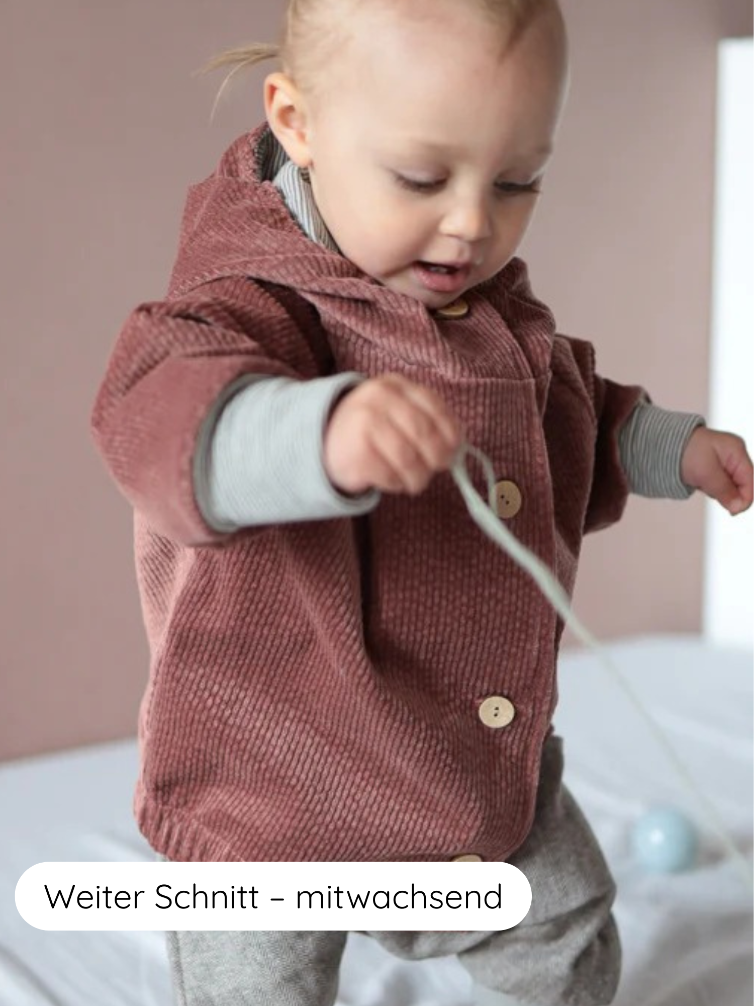 Kleinkind trägt Wichteljacke Breitcord Stretch- Rosenholz mit hölzernen Knöpfen, über grau gestreiftem Langarmshirt, stehend auf weicher Oberfläche, hält Schnur mit blauem Ball.