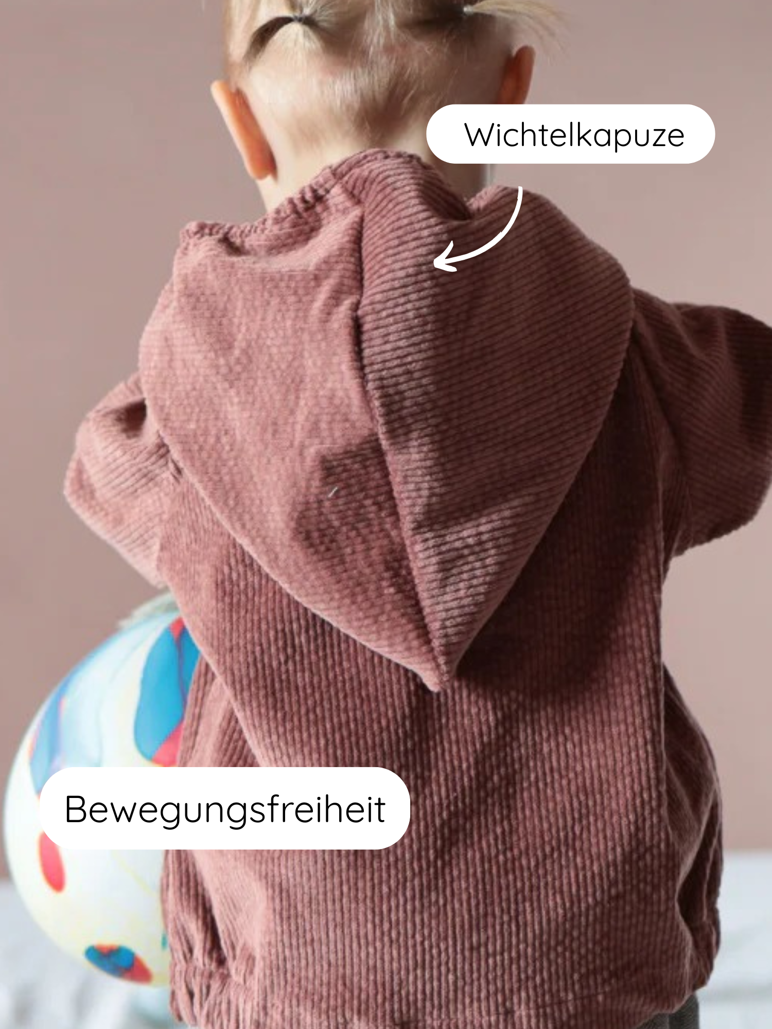 Ein Kind in der Wichteljacke Breitcord Stretch- Rosenholz mit spitzer Wichtelkapuze und breiten Krempel-Armbündchen aus Ringeljersey, hält einen bunten Ball, illustriert Bewegungsfreiheit und Komfort.