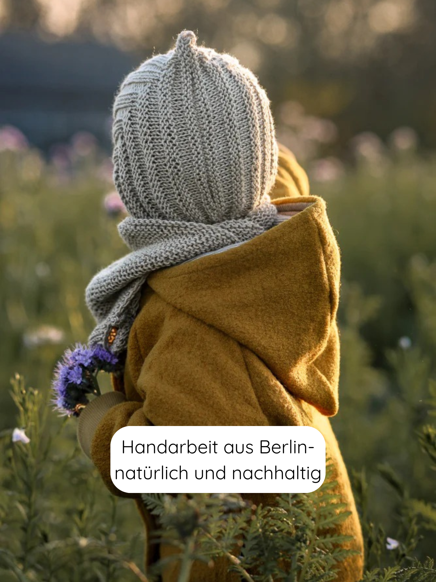 Ein Kind trägt eine Wichteljacke Wollwalk - Honig, gestrickte Balaklava und Schal in einem Wildblumenfeld. Die Jacke bietet Komfort und Bewegungsfreiheit, ideal für Herbst, Winter und Frühling.
