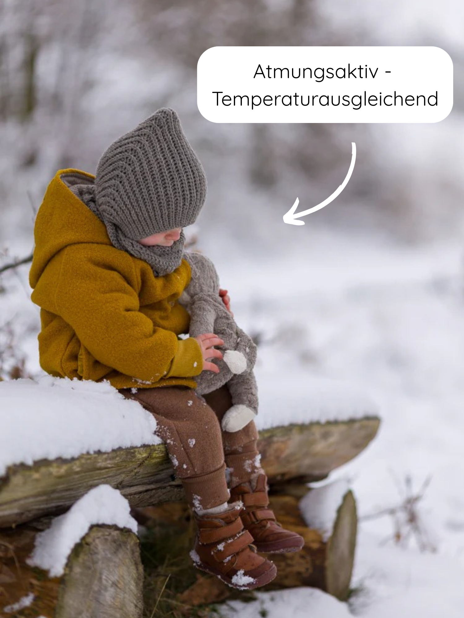 Kleinkind im Wollwalk-Wichteljacke - Honig, sitzt auf verschneitem Baumstamm, trägt braune Hosen, graue Mütze und Schal, hält graues Stoffkaninchen, mit Hinweis auf atmungsaktive, temperaturausgleichende Eigenschaften.