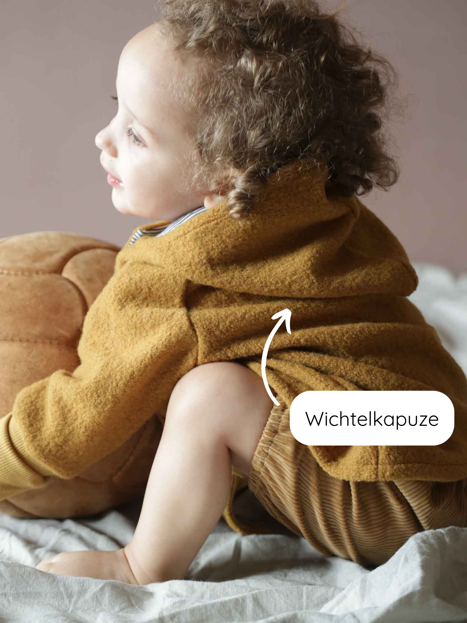 Kleinkind in einer Wichteljacke Wollwalk - Honig mit spitzer Kapuze, sitzt auf einer hellen Decke. Die Jacke ist warm, gefüttert und wächst mit, ideal für kühle Jahreszeiten.