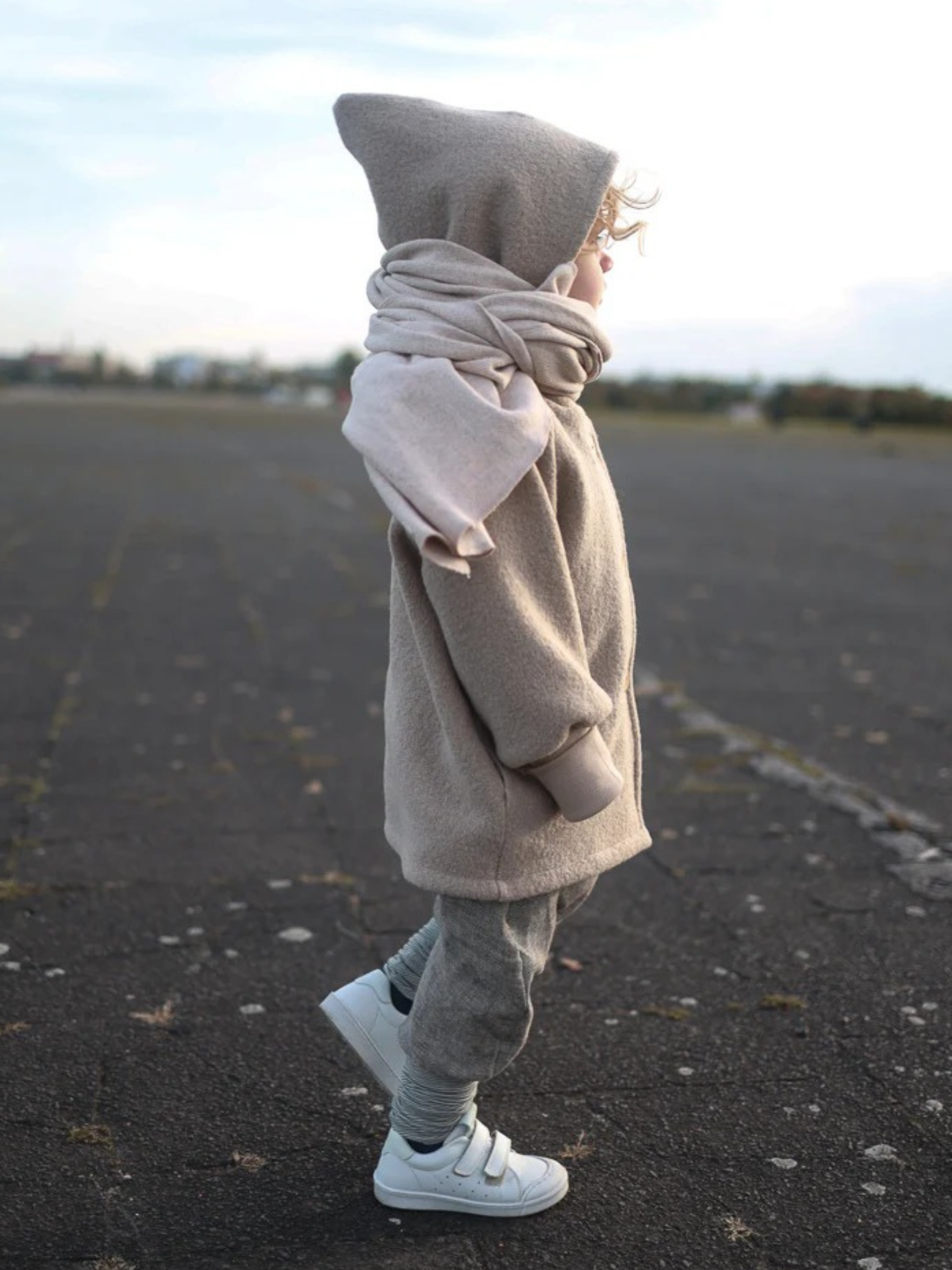 Ein Kind trägt die Wichteljacke Wollwalk - Sand von PETIT COCHON, eine beige, übergroße Kapuzenjacke mit grauen Jogginghosen und weißen Sneakers, auf einem leeren, mit Blättern bedeckten Asphalt.