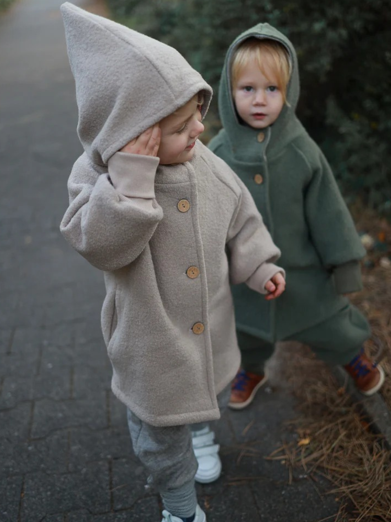 Zwei Kinder auf einem Weg, das vordere trägt die Wichteljacke Wollwalk - Sand mit Kapuze und Knöpfen, kombiniert mit grauer Hose und weißen Sneakers. Das hintere Kind trägt eine grüne Jacke.