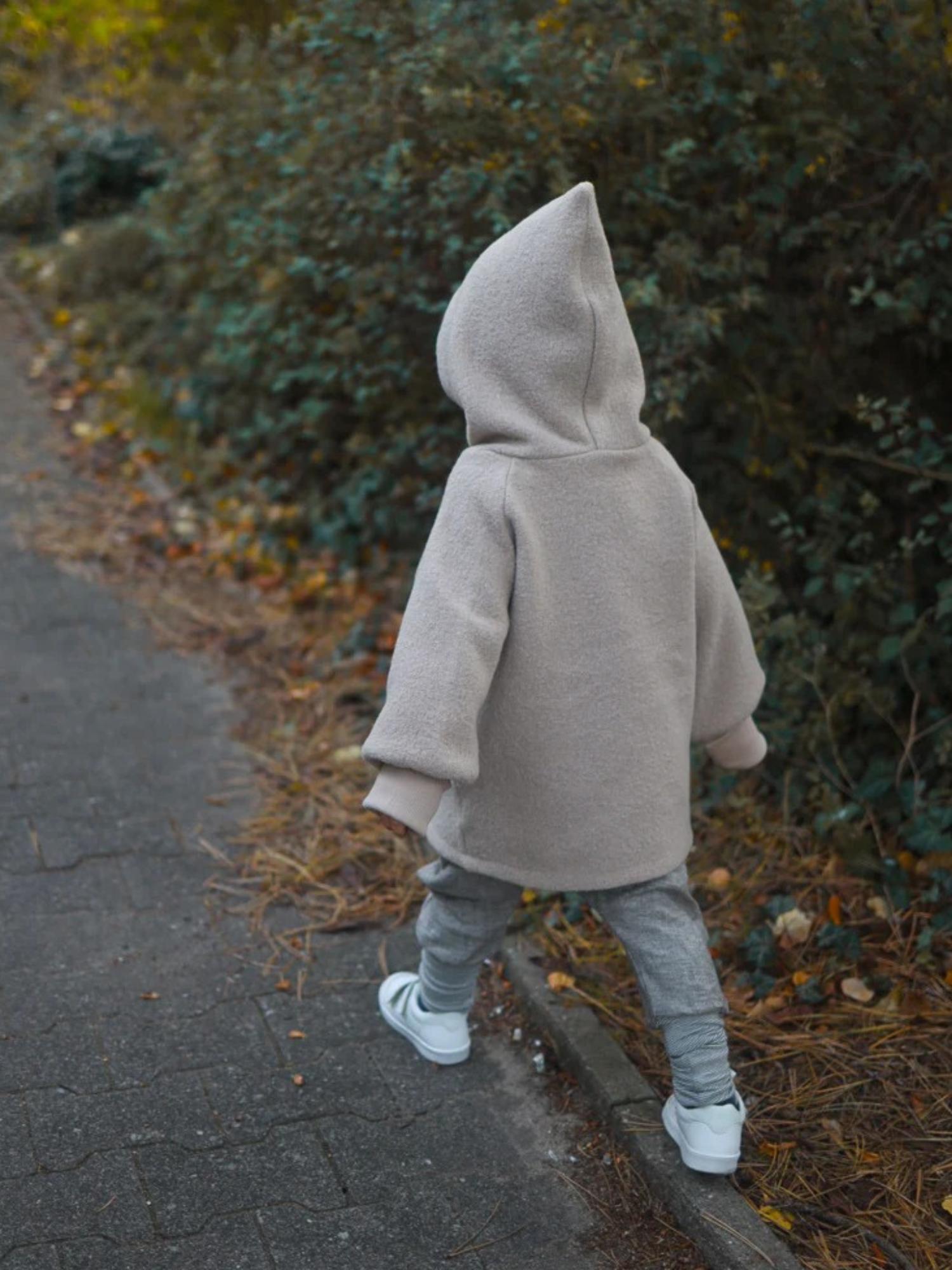 Ein Kind trägt die Wichteljacke Wollwalk - Sand, eine warme, graue Kapuzenjacke, auf einem herbstlichen Pfad spazierend. Die Jacke bietet Bewegungsfreiheit und ist ideal für kühle Jahreszeiten.