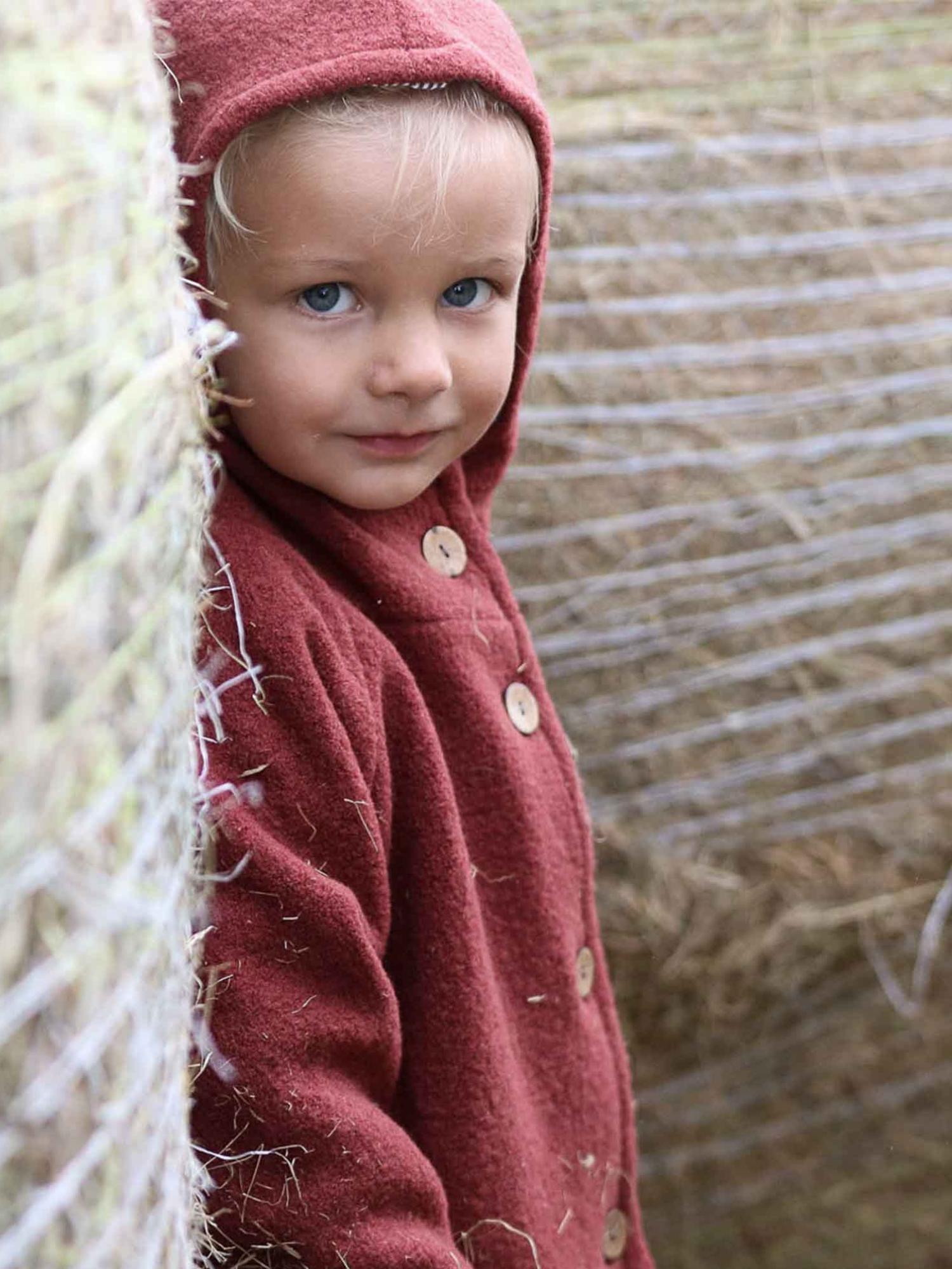 Ein Kleinkind trägt die Wichteljacke Wollwalk - Ziegel, eine rotgefärbte Wolljacke mit Holzknöpfen, während es neben großen Heuballen steht, ideal für kühle Jahreszeiten.