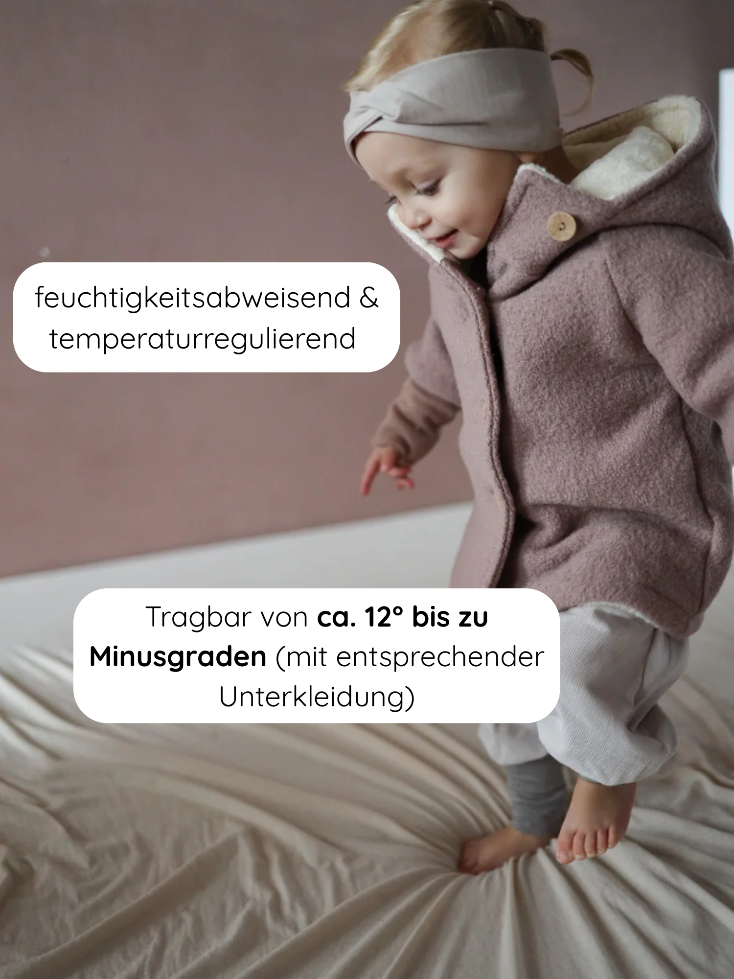 Kleinkind trägt die Winter Wichteljacke Wollwalk - Puder, steht auf einem weichen Bett; die Jacke hat eine spitze Kapuze, breite Bündchen und ein gemütliches Teddyfutter.