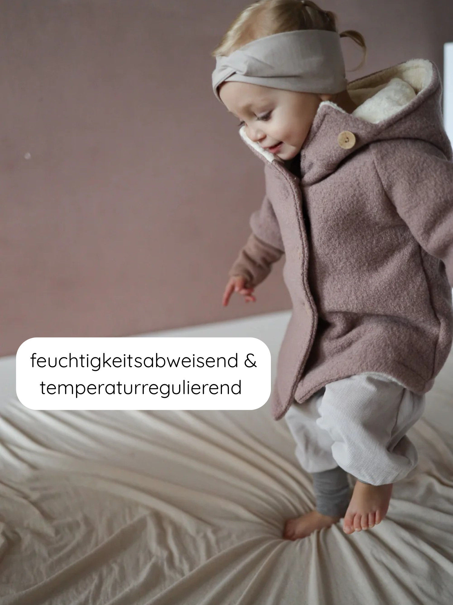 Kleinkind in Teddy-Wichteljacke Wollwalk - Puder, mit spitzer Kapuze und breiten Bündchen, sitzt auf einem Bett. Text betont die Feuchtigkeitsabweisung und Temperaturregulierung der Bettwäsche.