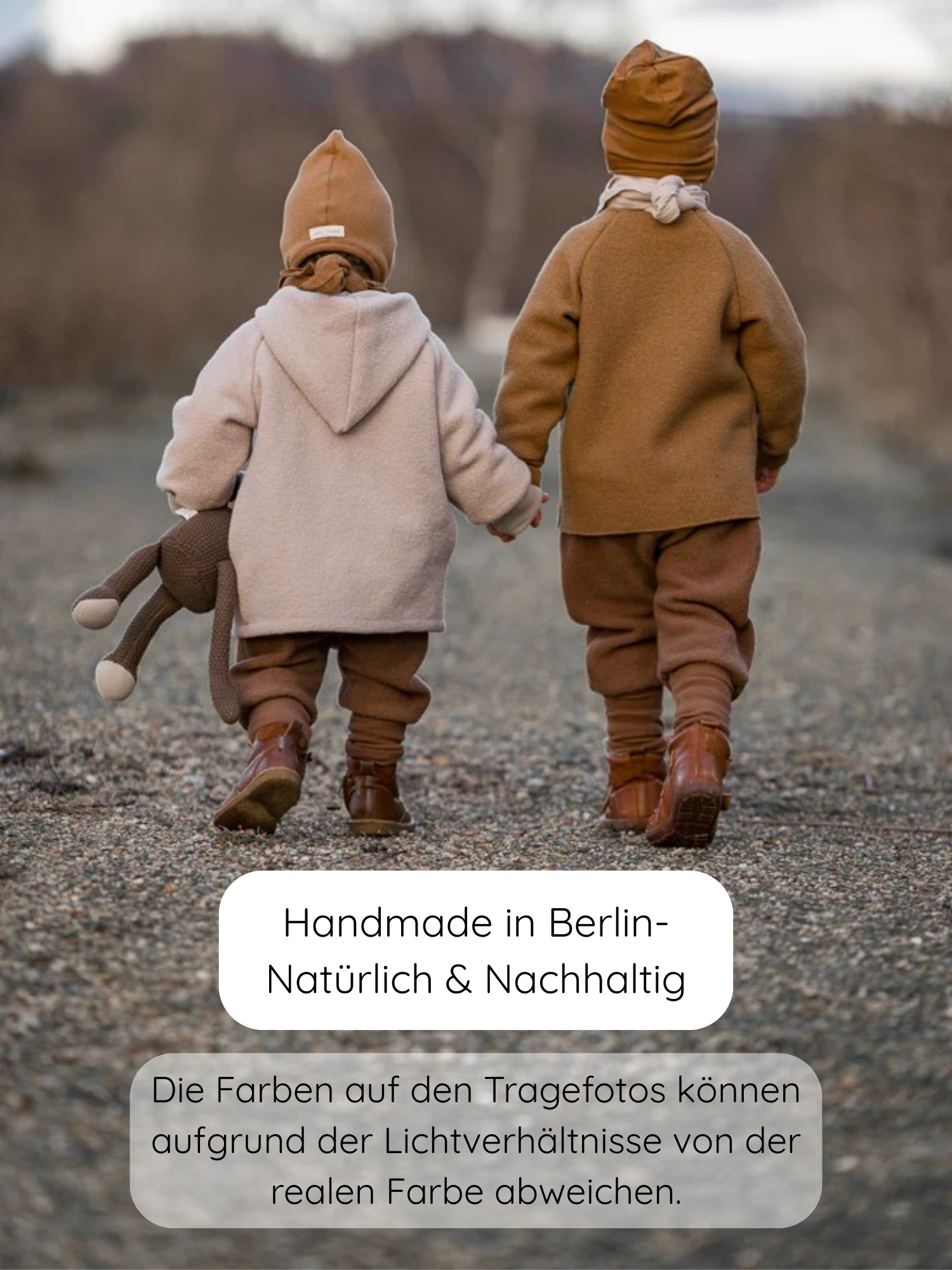 Zwei Kleinkinder in erdfarbenen Winter Wichteljacken Wollwalk - Sand, handgefertigt in Berlin, gehen Hand in Hand auf einem Kiesweg. Eines trägt ein gestricktes Kuscheltier.