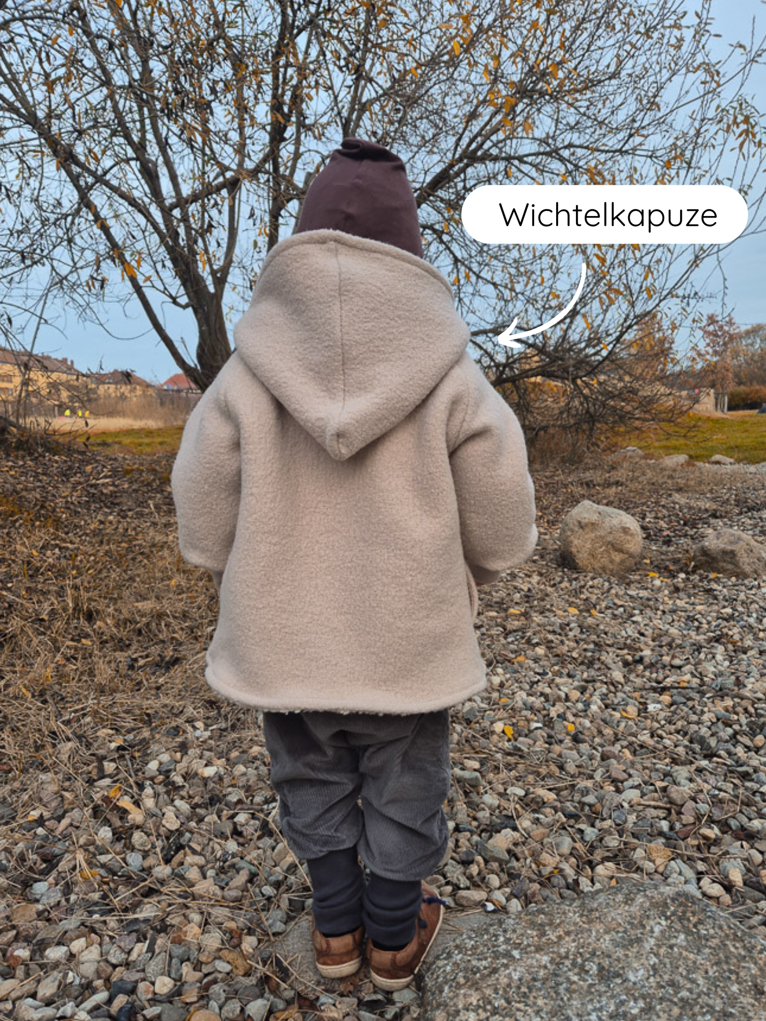 Ein kleines Kind trägt eine Winter Wichteljacke Wollwalk - Sand mit spitzer Kapuze, kombiniert mit burgunder Mütze, grauen Cordhosen und braunen Lederschuhen. Ideal für kalte Tage.