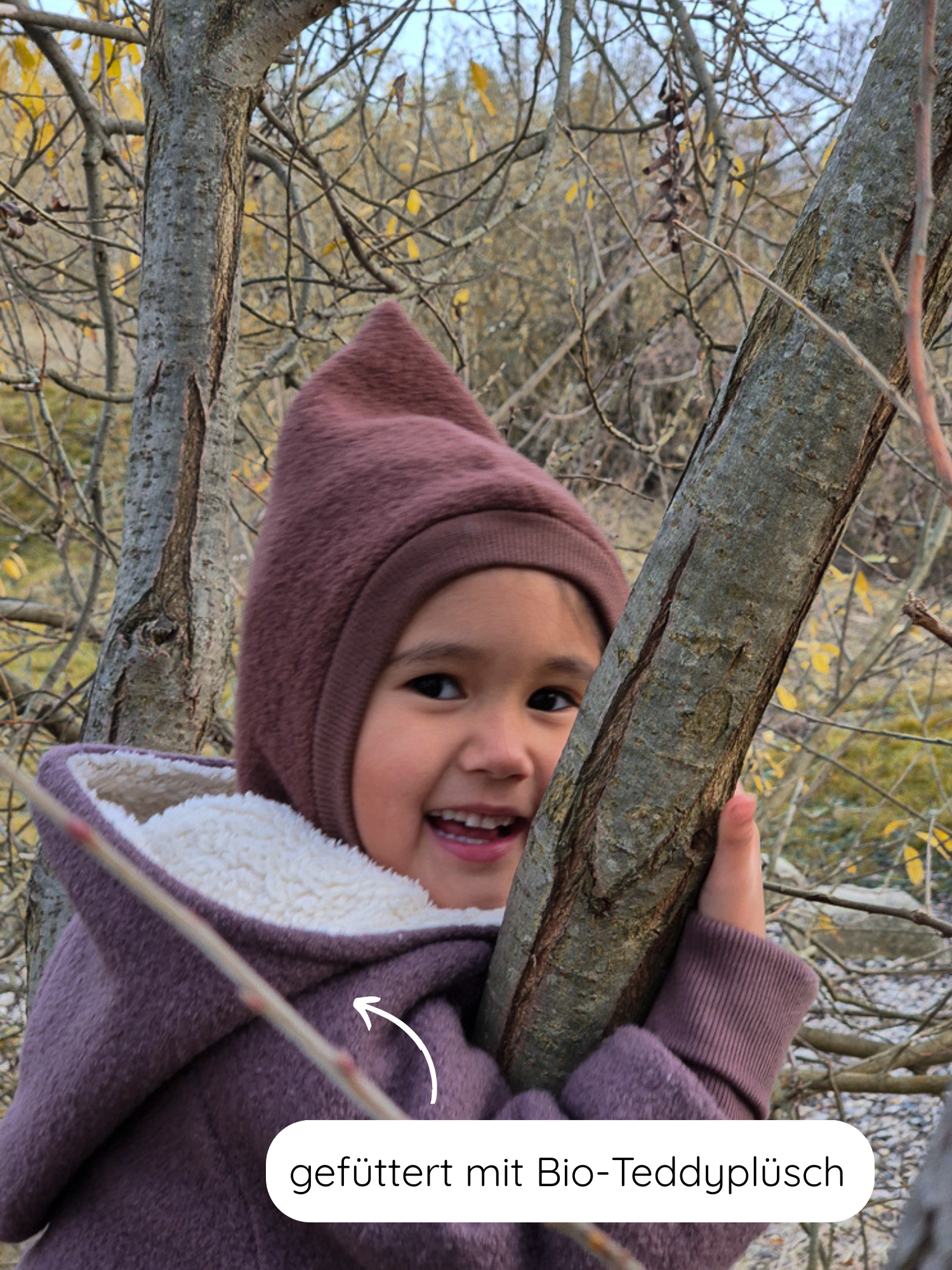 Kind in einem parkähnlichen Umfeld, trägt die Winter Wichteljacke Wollwalk mit Teddyfutter - Malve. Die Jacke hat eine spitze Kapuze und weiche, gefütterte Innenseite aus Bio-Baumwolle.