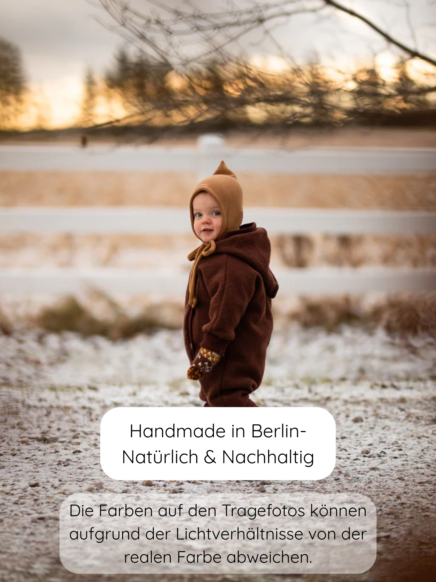 Kleinkind trägt den Kuschelanzug Wollwalk - Schoko mit spitzer Kapuze und passenden Fäustlingen, stehend im Schnee vor einem weißen Holzzaun. Handgefertigt in Berlin, umweltfreundlich und nachhaltig.