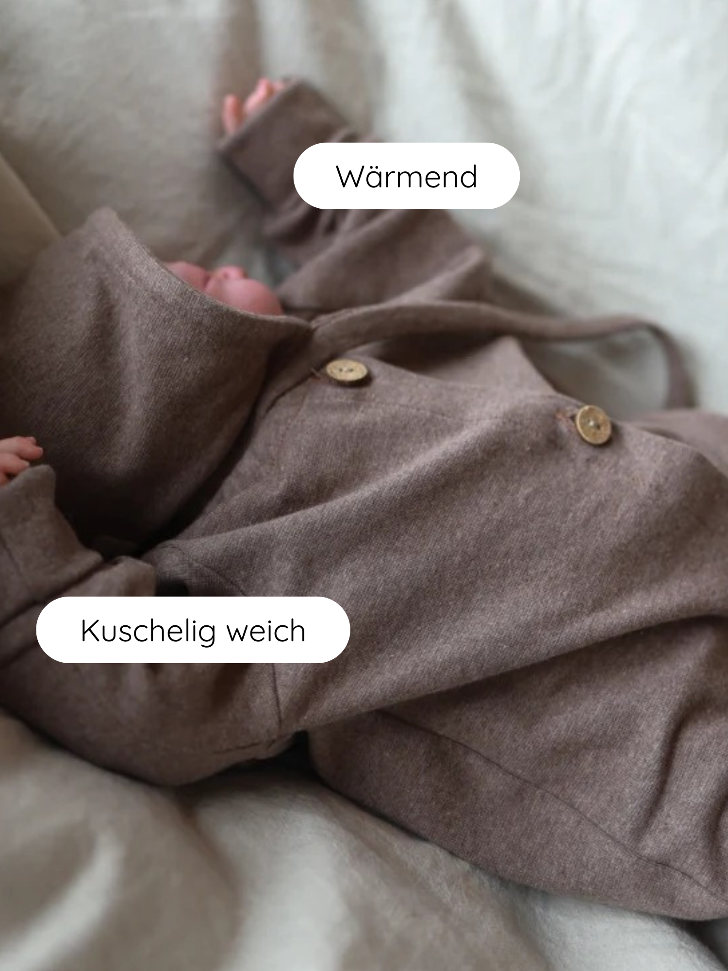 Ein Baby trägt die Zwergenmütze Feinstrick - Praline Meliert, eine warme, braune Kapuzen-Einteiler mit Daumenlöchern und Holzknöpfen, auf einem weichen, hellen Untergrund liegend.