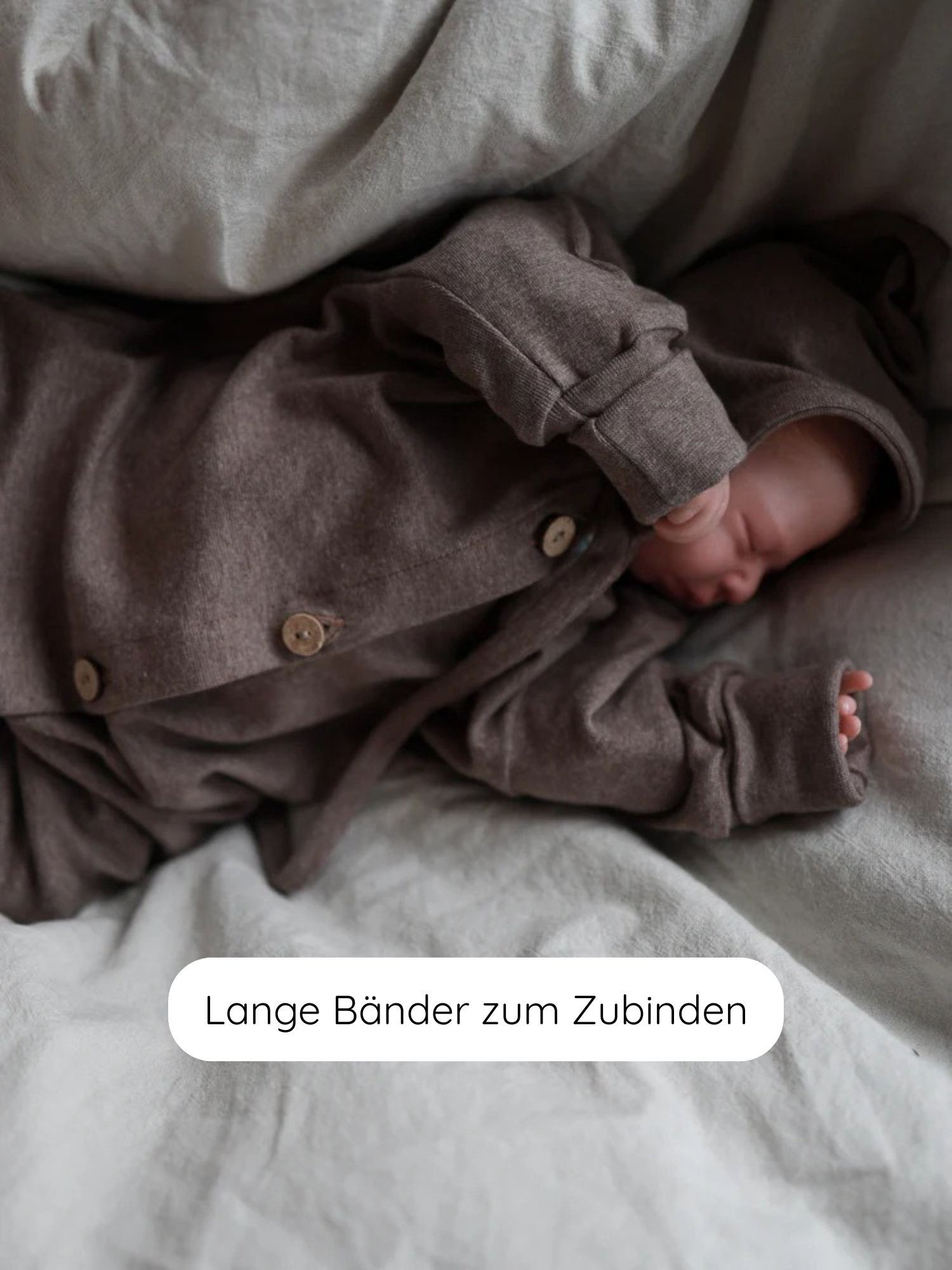 Schlafendes Neugeborenes in Zwergenmütze Feinstrick - Praline Meliert mit langen Bändern zum Zubinden, auf hellem Bett, zeigt die weiche, elastische Struktur des melierten Jersey-Stoffes.