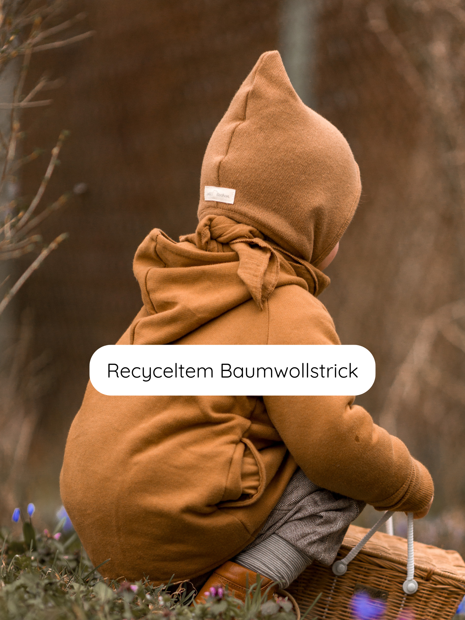 Kleinkind von hinten in warmer Strickkleidung mit Zwergenmütze Strick - Sahara aus recycelter Baumwolle, hält einen Korb im Frühlingsgras.