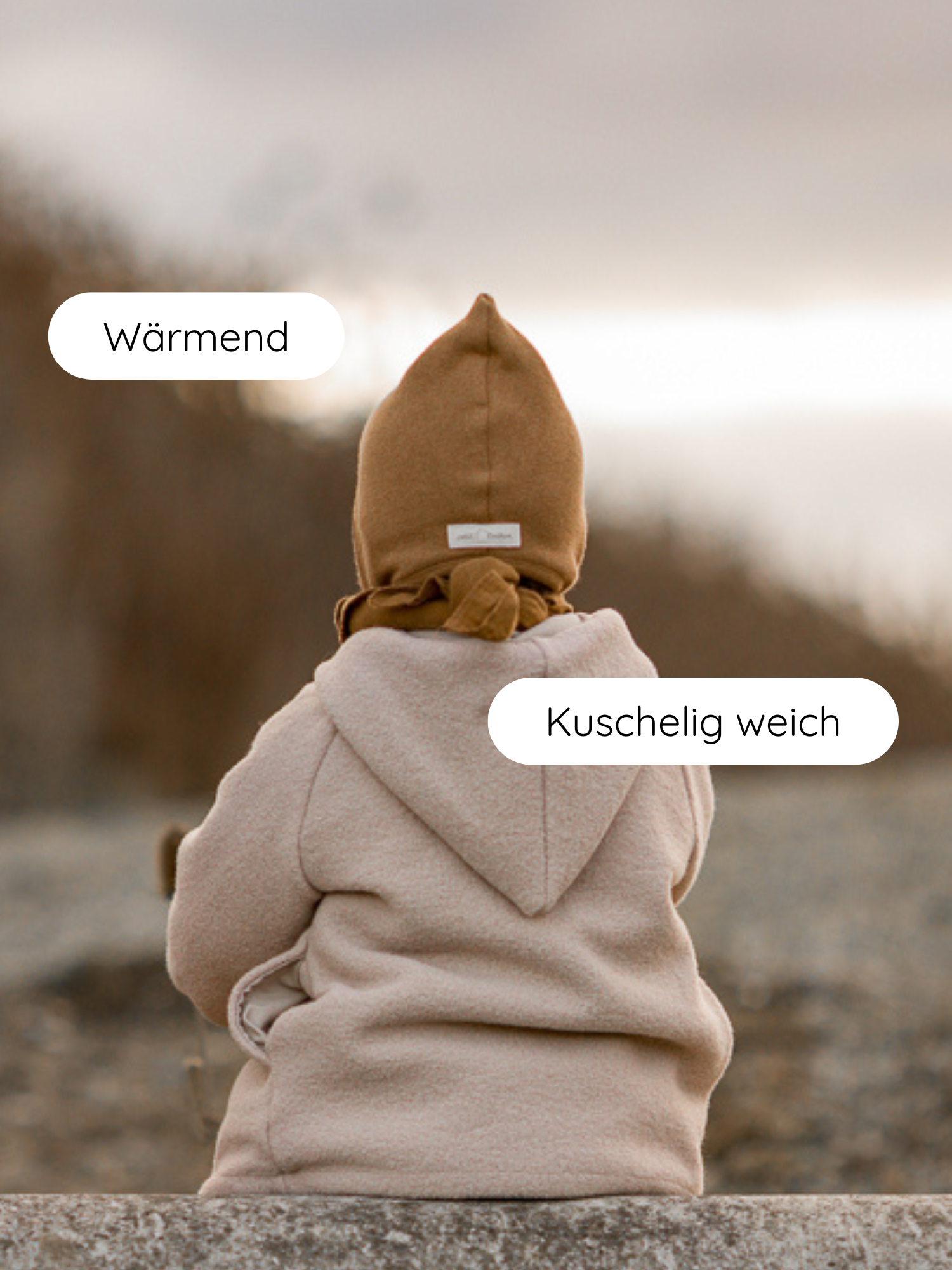 Ein Kind trägt die Zwergenmütze Strick - Sahara aus recycelter Baumwolle, sitzt am felsigen Ufer. Der warme, weiche Mütze sorgt für Komfort und Schutz in kühler Umgebung.