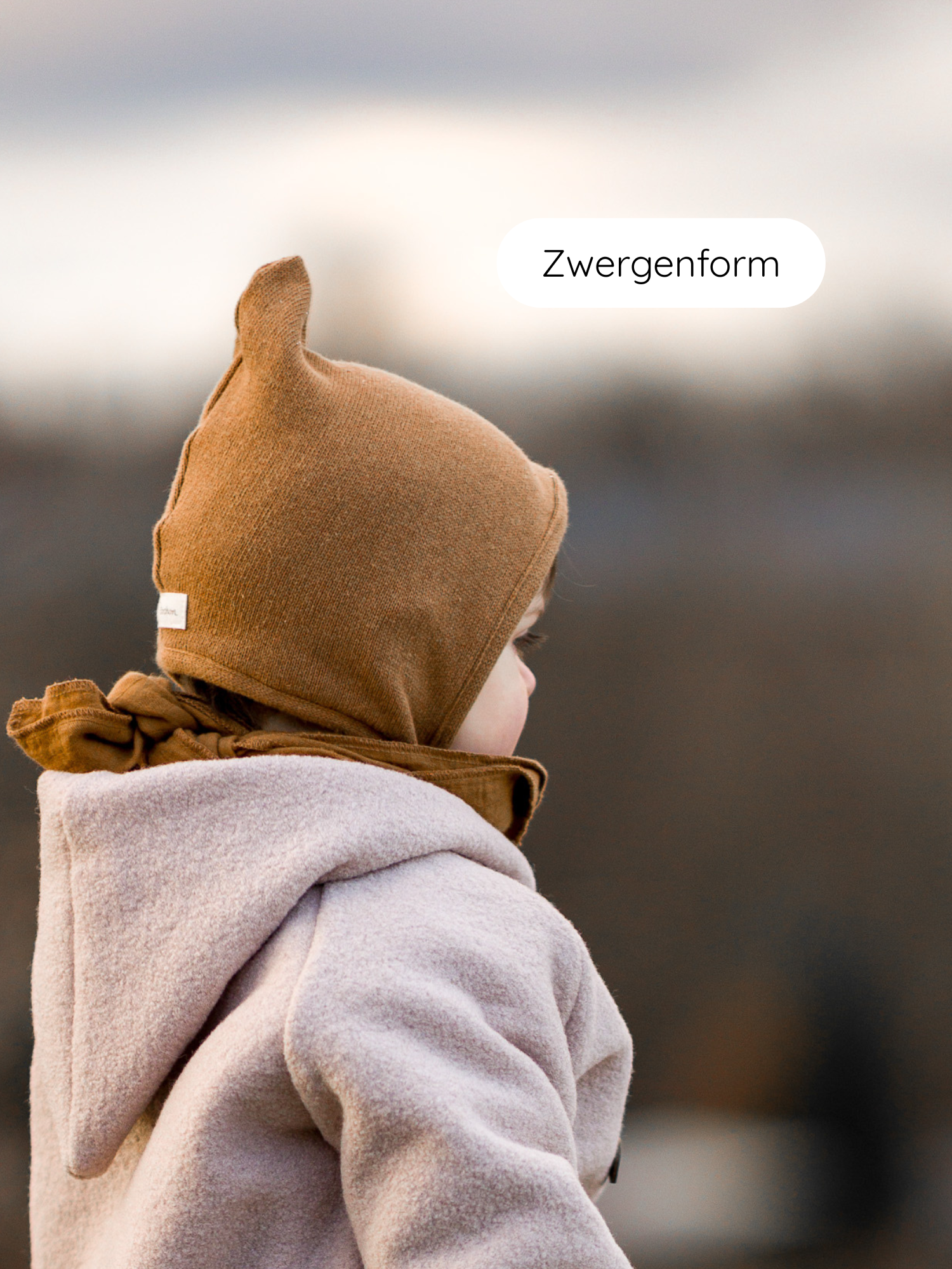 Kind in Zwergenmütze Strick - Sahara aus recyceltem Baumwollstrick, seitlich fotografiert, mit spitzem, braunem Bonnet, weichem Fleece-Mantel, draußen im unscharfen Hintergrund, beschriftet mit Zwergenform.