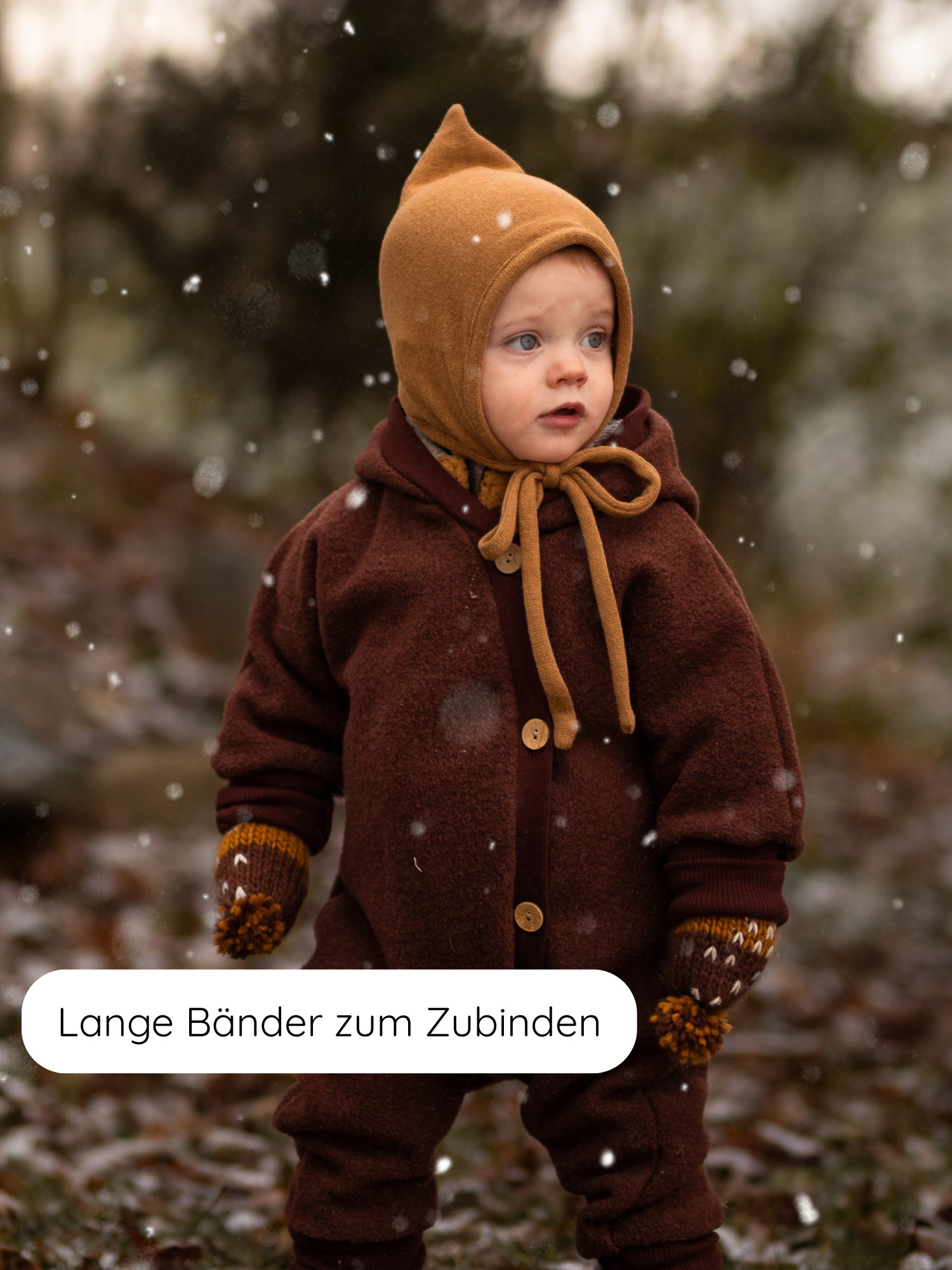 Kleinkind im leichten Schneefall trägt die Zwergenmütze Strick - Sahara, ein warmes, recyceltes Baumwollaccessoire mit langem Bindeband, passend zu seinem braunen Einteiler und gelben Strickhandschuhen.