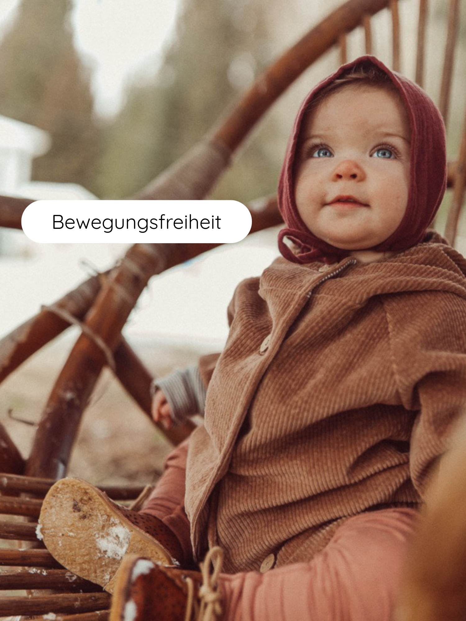 Kleinkind in braunem Babyknicker Kaschmir-Mix mit Klappbündchen, sitzend auf einem rustikalen Stuhl. Das Outfit umfasst eine Kapuze, tiefrote Haube und weiche Stiefel mit Schneeresten.