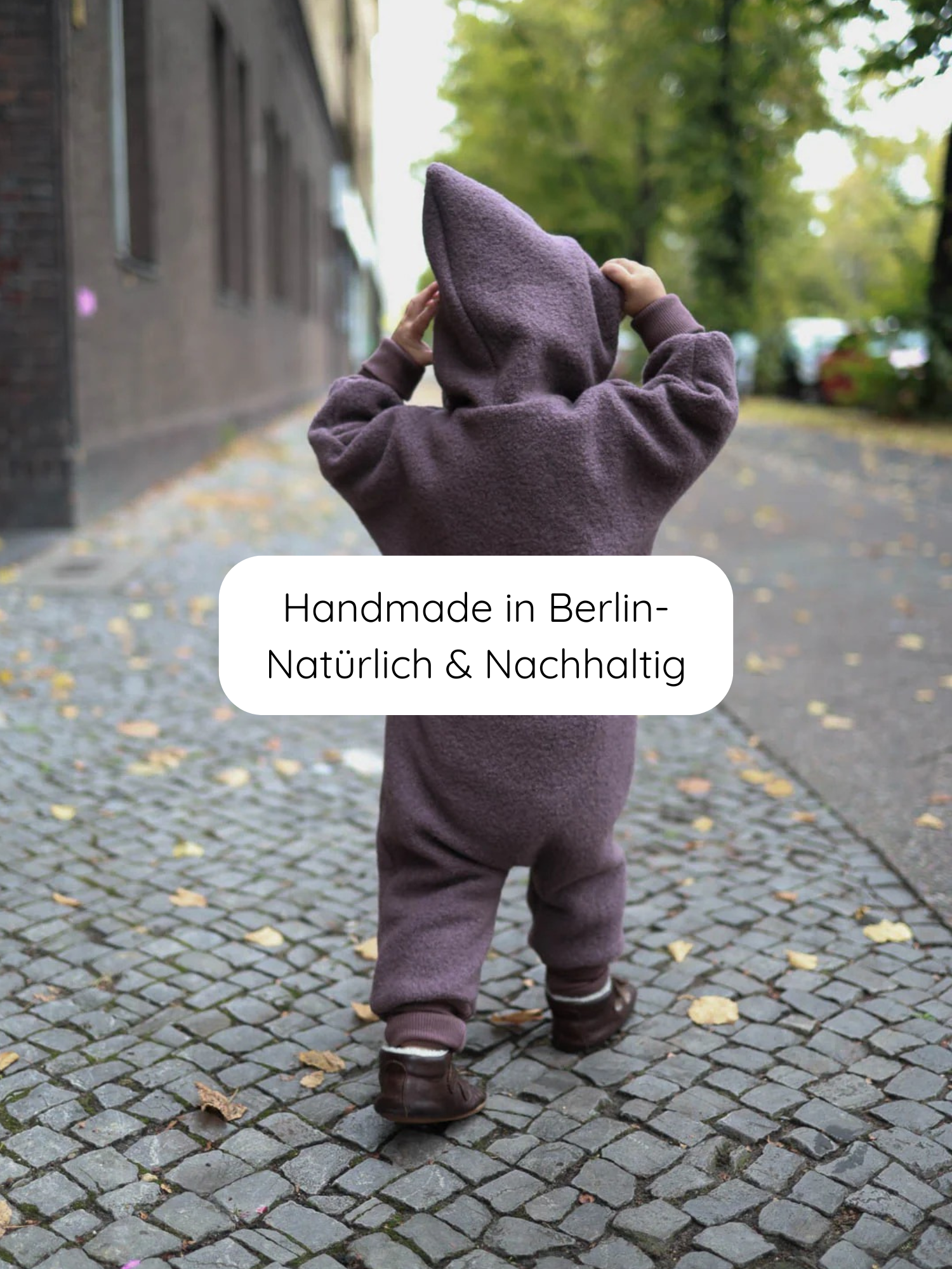 Kleinkind von hinten auf Kopfsteinpflaster, trägt den Kuschelanzug Wollwalk - Malve, einen lila Einteiler mit Kapuze. Text: “Handmade in Berlin – Natürlich & Nachhaltig.”