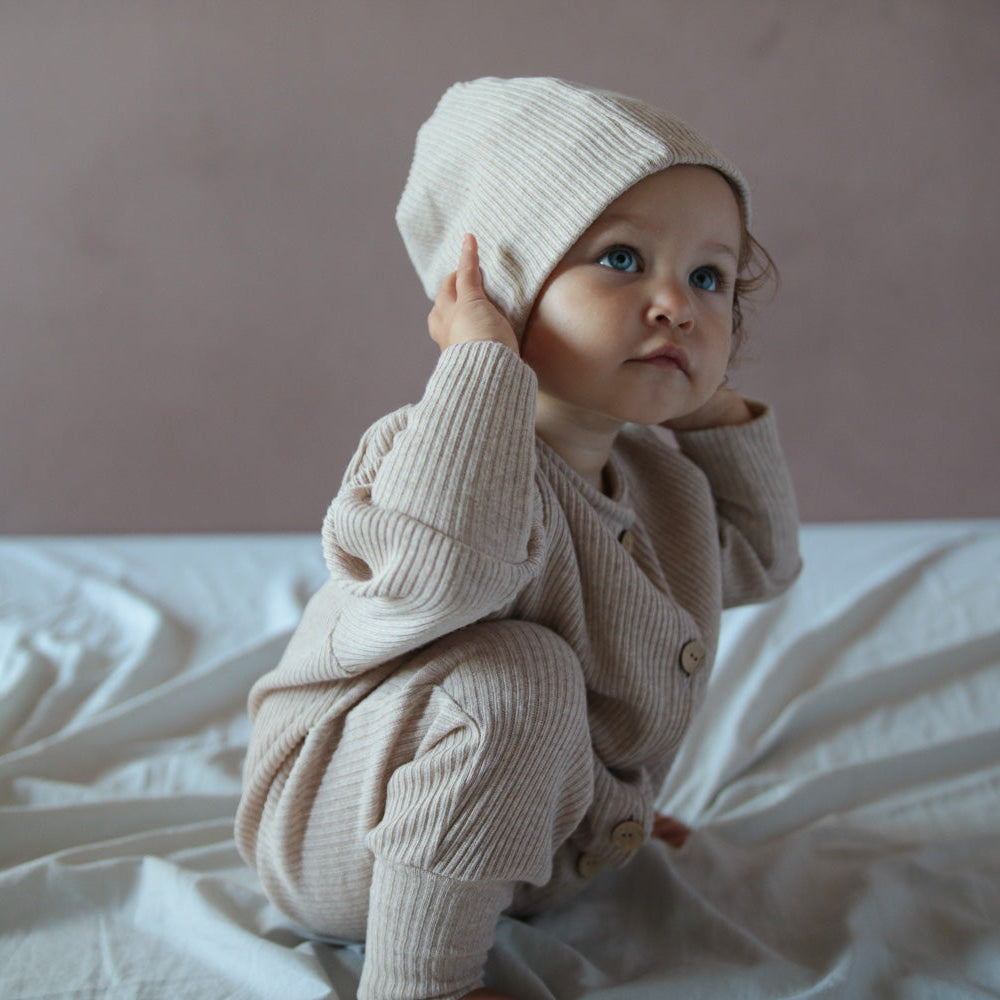 Kind sitzt auf dem Bett. Trägt Strampler und Beanie. Nachhaltige Kinderkleidung und Baby Erstausstattung von Petit Cochon.
