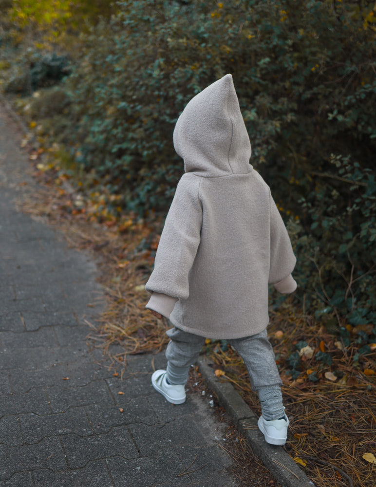 Ein kleines Kind in einer leicht übergroßen Wichteljacke Wollwalk - Sand, aus Wollwalk mit Bio-Jersey-Futter, geht auf einem Weg entlang, passend für Herbst, Frühling und Winter.