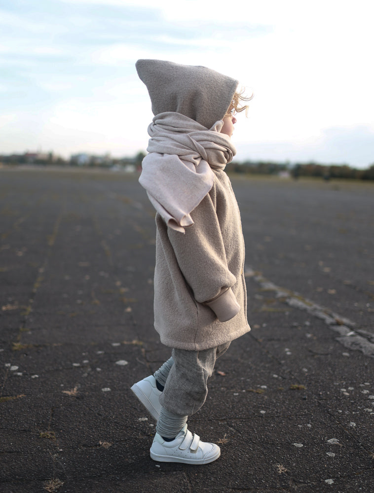 Kind seitlich stehend auf Asphaltfläche, trägt die Wichteljacke Wollwalk - Sand von PETIT COCHON. Die Jacke hat einen weichen, mitwachsenden Schnitt, krempelbare Bündchen und bietet große Bewegungsfreiheit.