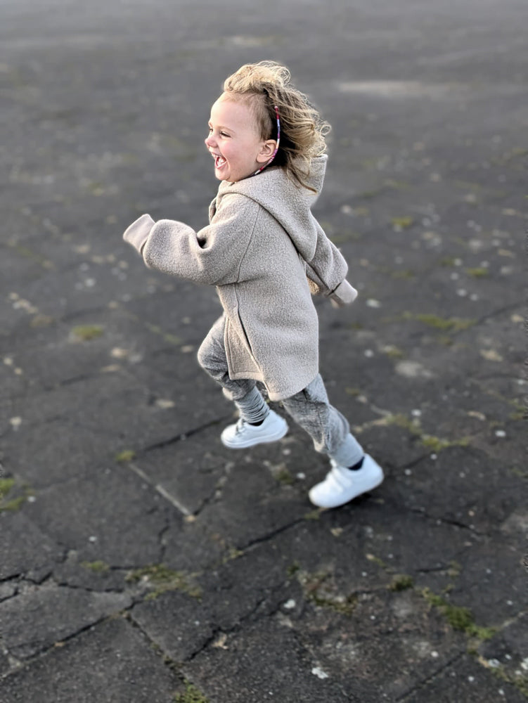 Ein Kind rennt lachend in der Wichteljacke Wollwalk - Sand über rissigen Asphalt. Die Jacke bietet Bewegungsfreiheit und Komfort, ideal für kühles Wetter.