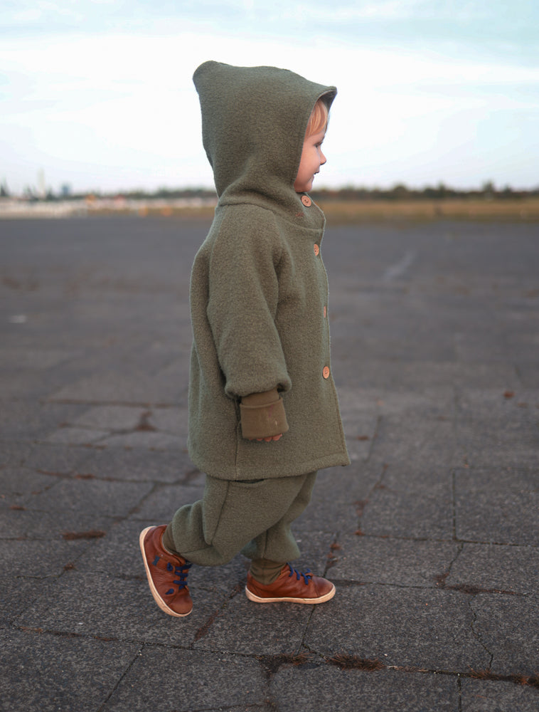 Ein Kleinkind in Knickerbocker Wollwalk - Pistazie, gehend auf breiter Fläche, trägt eine olivgrüne Fleecejacke und braune Ledersneaker mit blauen Schnürsenkeln.