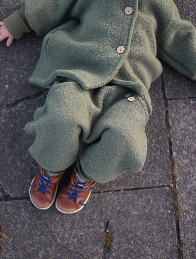 Kleinkind trägt Knickerbocker Wollwalk - Pistazie, aus olivgrünem Fleece mit Holzknöpfen. Kombiniert mit braunen Sneakers und blauen Schnürsenkeln, sitzt es auf grauen Fliesen.