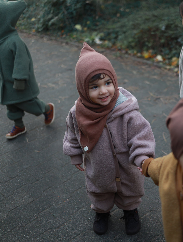 Kind im Kuschelanzug Wollwalk - Puder, aus 100% Schurwolle mit Baumwollfutter, auf einem Gehweg. Der Anzug bietet Bewegungsfreiheit und wärmt ideal für Herbst, gehalten von einer Hand.
