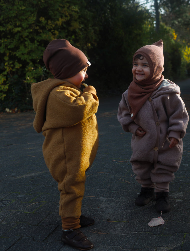 Zwei Kleinkinder im Kuschelanzug Wollwalk - Puder stehen auf einem Weg. Der Anzug bietet Bewegungsfreiheit und Schutz, ideal für kühle Spaziergänge, mit Bio-Baumwollbündchen und Futter.