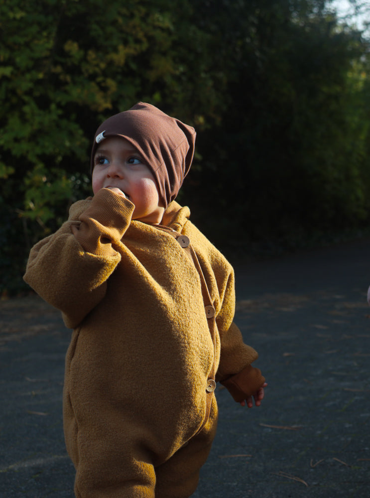 Ein Kleinkind trägt den Kuschelanzug Wollwalk - Honig aus 100% Schurwolle mit Holzknöpfen, während es auf einem Weg steht. Es trägt dazu eine braune Mütze und schaut zur Seite.