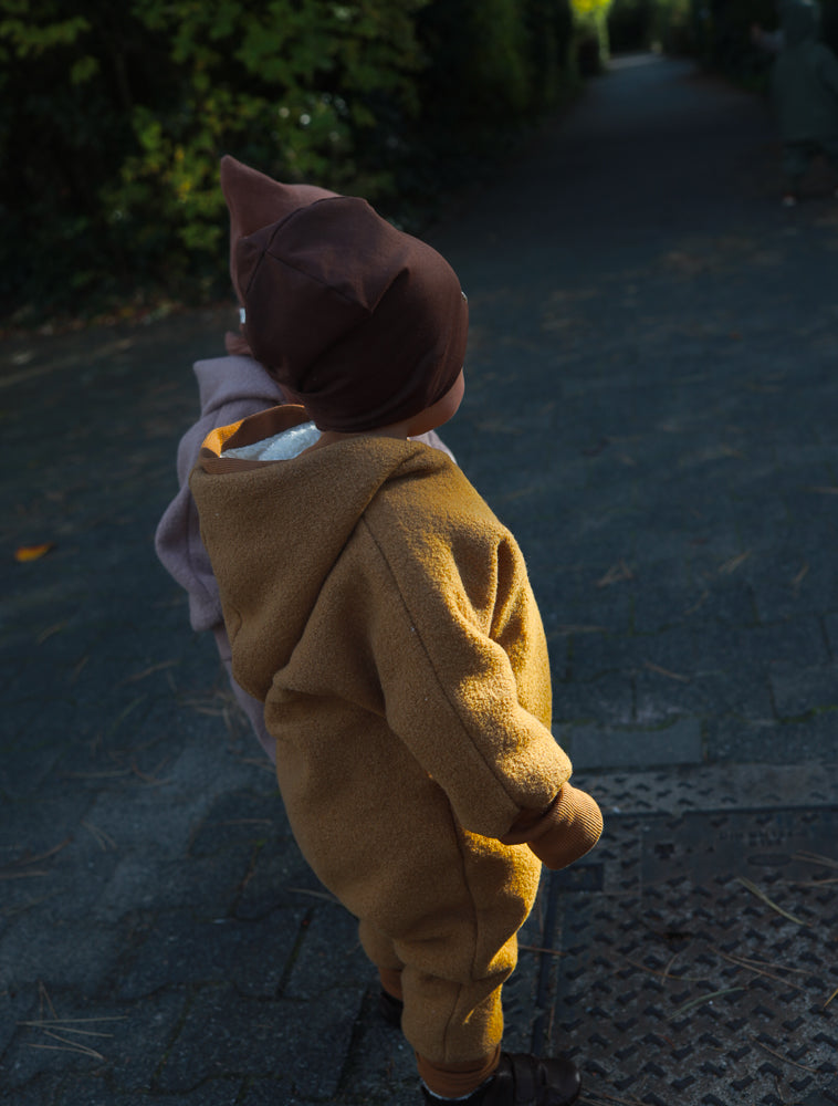 Kleinkind von hinten in einem Winter Wollwalk Overall mit Teddyfutter - Honig auf einem Gartenweg, umgeben von Bäumen, trägt eine dunkelbraune Mütze. Perfekt für kalte Wintertage.