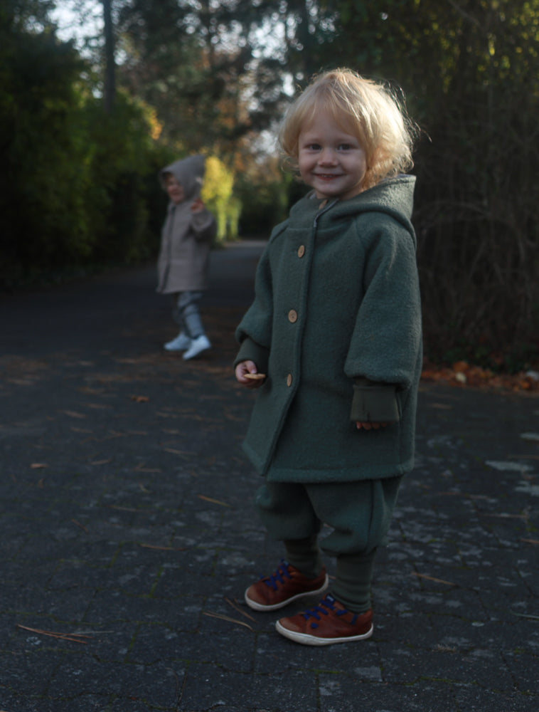 Zwei Kinder im Freien, im Vordergrund trägt eines die Knickerbocker Wollwalk - Pistazie mit Kängurutaschen, perfekt für kühles Wetter, geeignet für Spielplatz und Waldkindergarten.