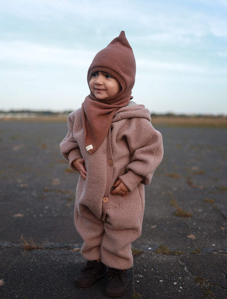Kleinkind im Kuschelanzug Wollwalk - Puder, einteilig mit Kapuze und integriertem Schal, aus weicher Schurwolle, trägt dunkle Stiefel auf gepflastertem Untergrund, Hintergrund unscharf. Ideal für kühlere Tage.
