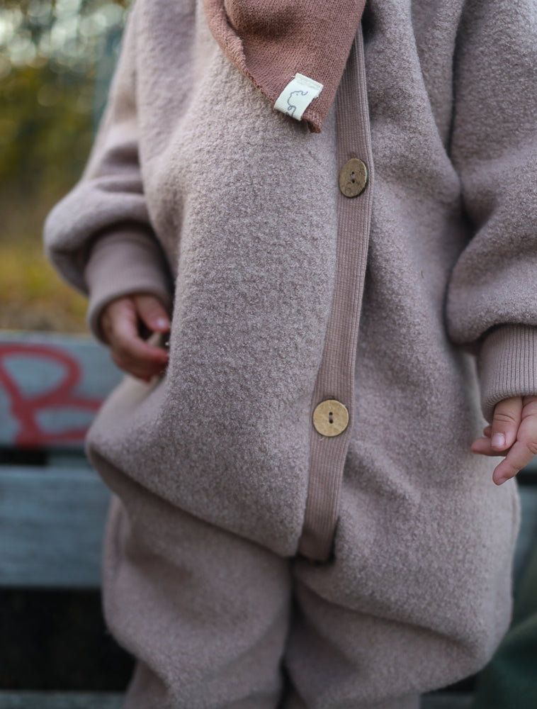 Kuschelanzug Wollwalk - Puder: Kind im weichen, beigen Zweiteiler aus Fleece mit Knopfjacke und passender Hose, ergänzt durch ein leichtes braunes Halstuch.