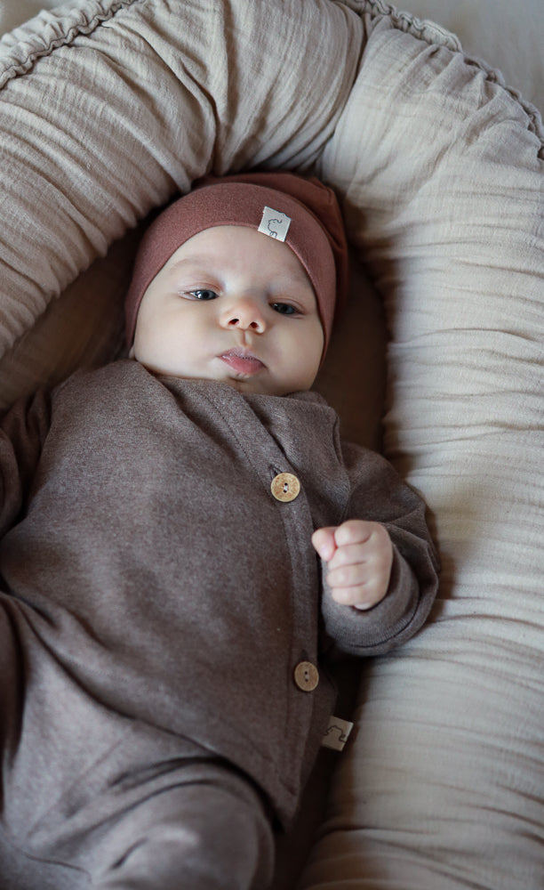 Ein Baby trägt den Babyschlüttli Feinstrick - Praline Meliert, einen braunen, weichen Jersey-Overall mit seitlichem Verschluss, kombiniert mit einer passenden Mütze, liegend in einem gepolsterten Baby-Lounger.