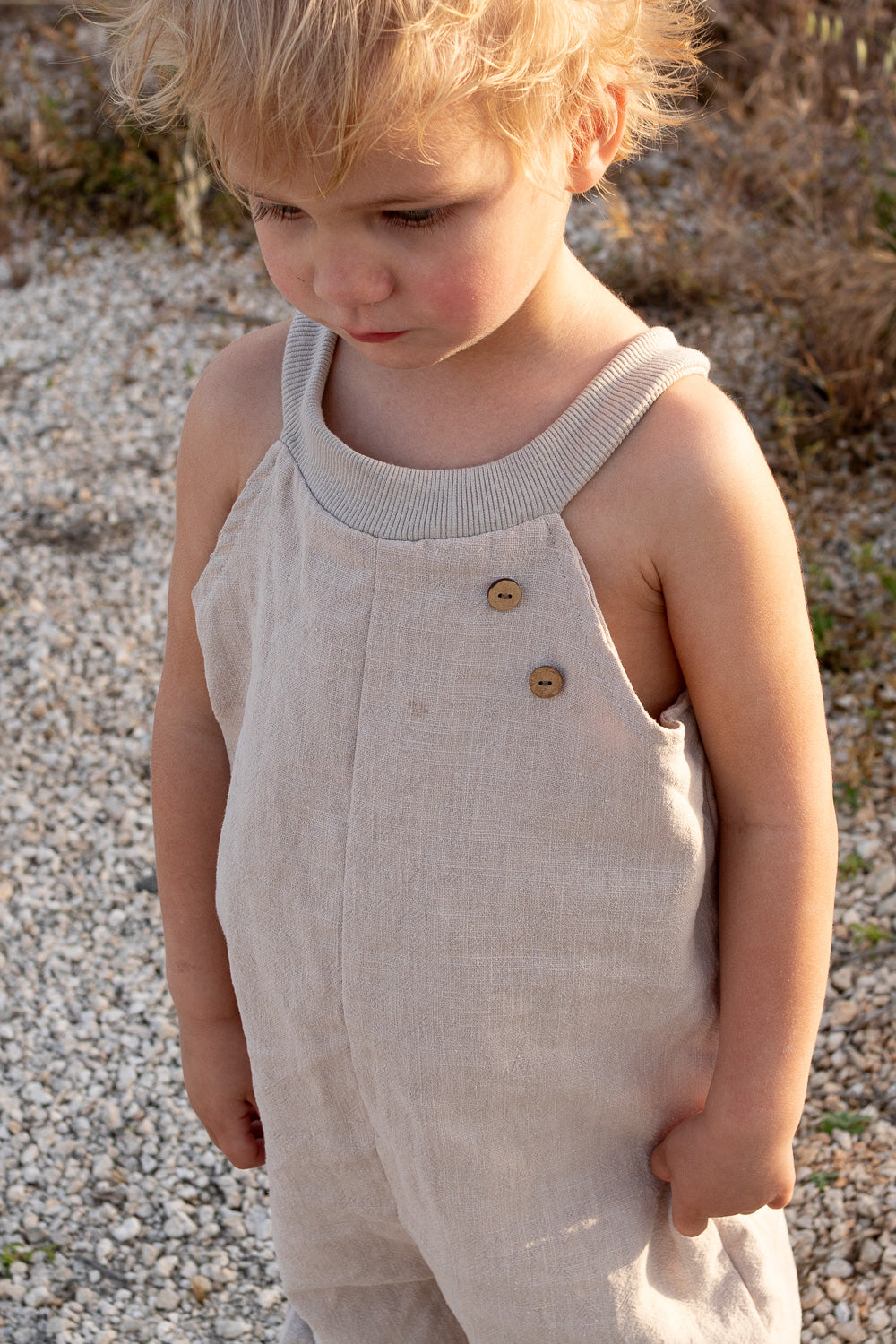 Kind trägt Schlupflatzhose Leinen - Sand, eine beige, lockere Leinenhose mit Holzknöpfen und geripptem Kragen. Ideal für Bewegungsfreiheit, aus atmungsaktivem Material, perfekt für Frühling und Sommer.