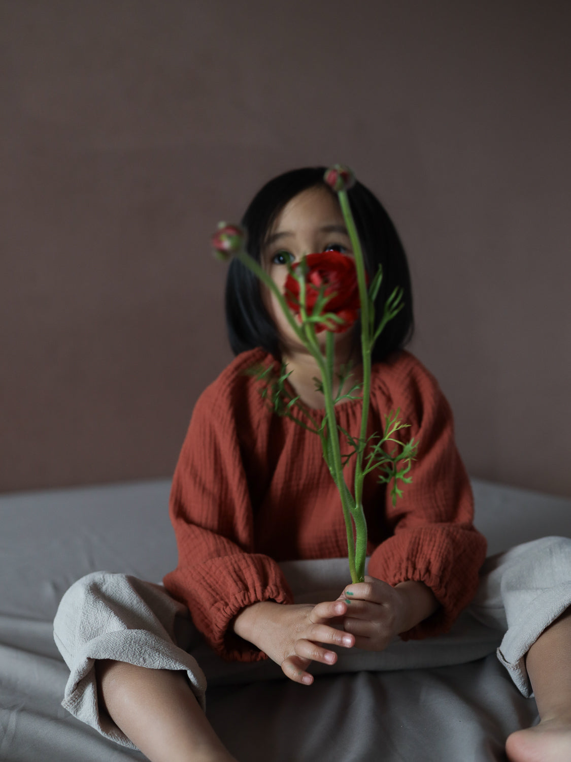 Ein Kind sitzt im Blüschen Musselin - Terrakotta, locker geschnitten, hält eine rote Blume, betont die luftige, weiche Struktur des Baumwoll-Musselins, ideal für warme Tage.
