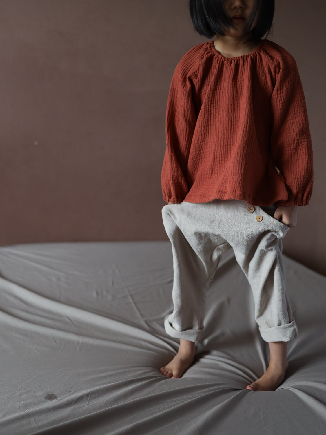 Kind trägt Blüschen Musselin - Terrakotta, locker und langärmelig, ideal für warme Tage. Kombiniert mit hellen, übergroßen Hosen mit Holzknöpfen. Leicht und angenehm für empfindliche Haut.