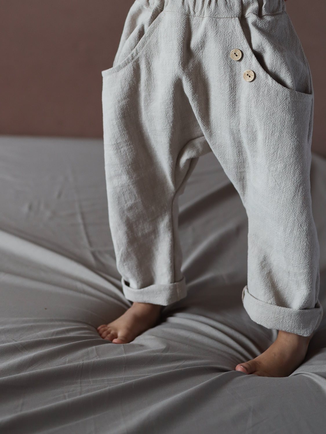 Kleinkind steht barfuß auf grauem Untergrund, trägt die Sommerknicker Leinen - Sand: locker geschnittene, beige Leinenhosen mit aufgerollten Bündchen und zwei Holzknöpfen an der Fronttasche.