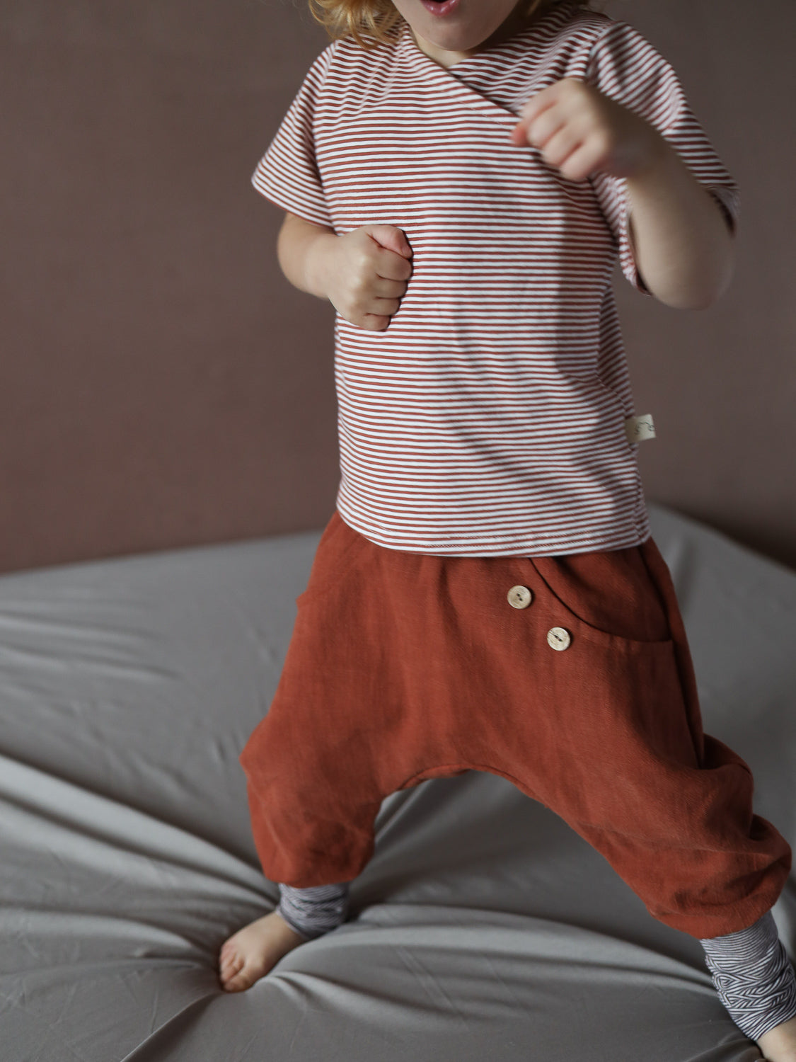 Kleinkind trägt ein kurzes Wickelshirt aus Baumwolle - Ringel Ziegel, mit schmalen Streifen, kombiniert mit weiten Hosen. Shirt ist bequem und lässig, ideal für sommerliche Outfits.