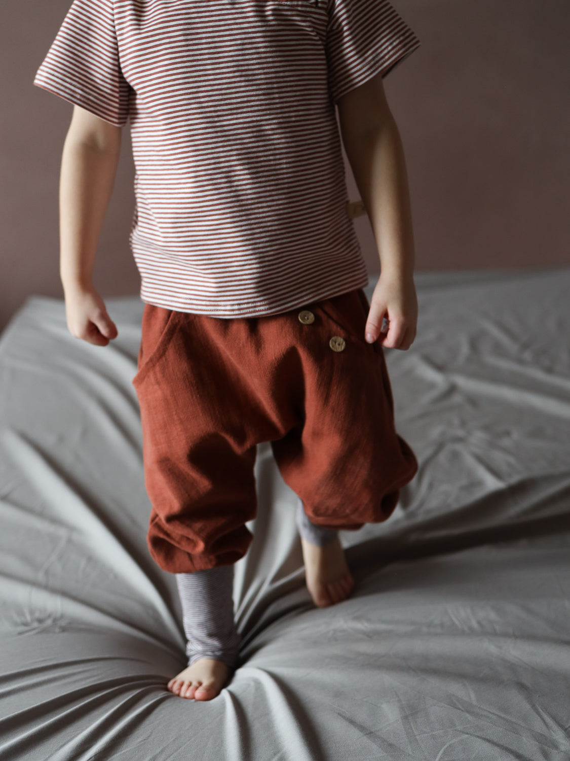 Ein Kind trägt das Kurzes Wickelshirt Baumwolle - Ringel Ziegel, ein kurzärmeliges, rot-weiß gestreiftes Shirt, kombiniert mit lockeren Hosen und gestreiften Beinwärmern. Perfekt für sommerliche Outfits.