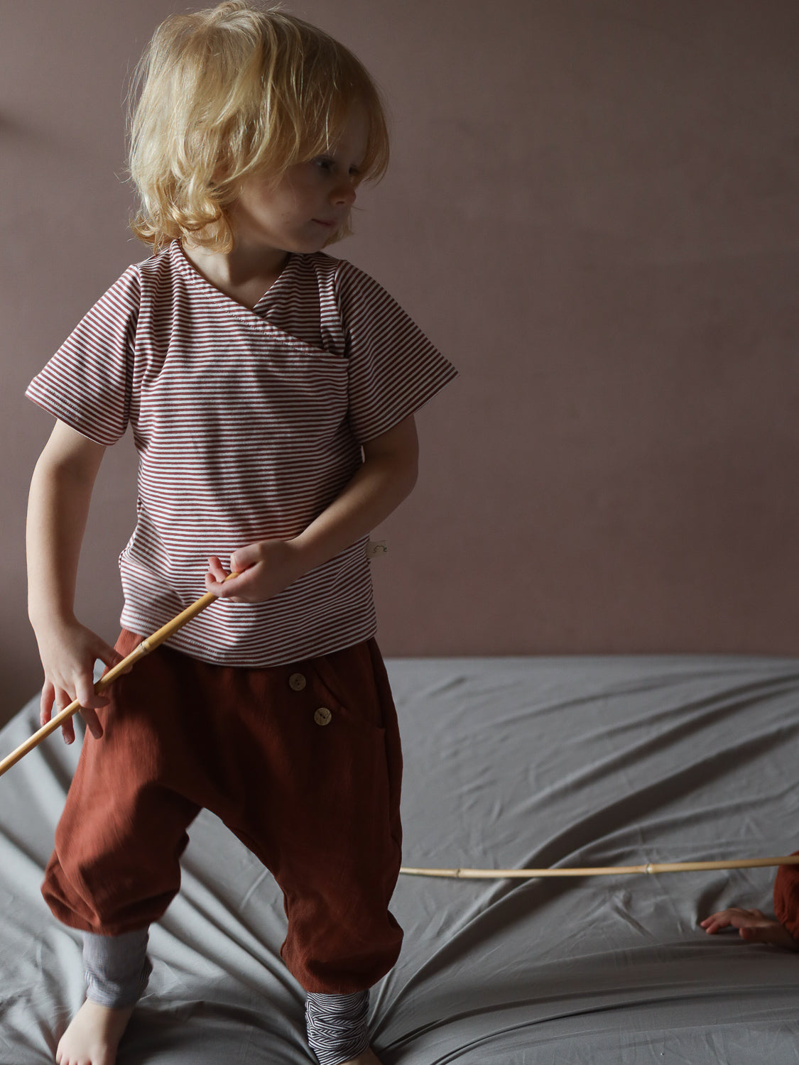 Kleinkind in kurzes Wickelshirt Baumwolle - Ringel Ziegel, mit feinen Streifen, hält einen Stock. Das Shirt ist kurzärmelig und kombiniert mit rustfarbenen Hosen.