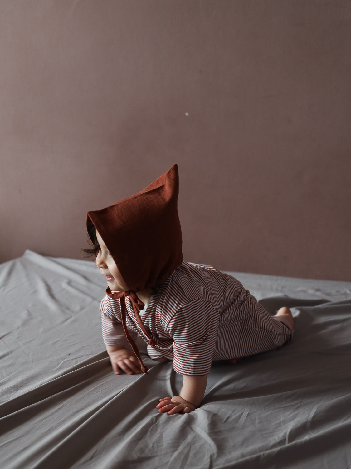 Baby in Wickel Overall Baumwolle - Ringel Ziegel auf weichem, grauem Laken, krabbelt auf Händen und Knien. Der Overall hat rot-weiße Streifen, lange Ärmel und eine bequeme Passform.