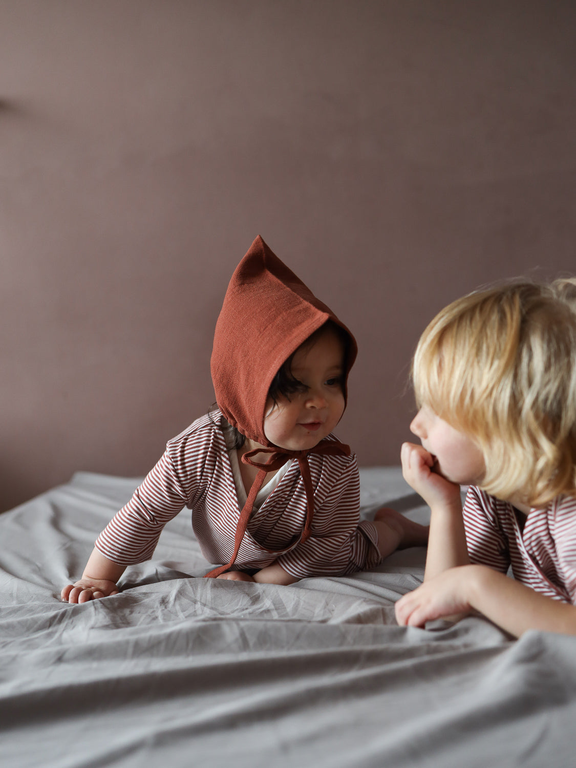 Zwei Kinder auf einem Bett, im Vordergrund ein Baby im Wickel Overall Baumwolle - Ringel Ziegel, auf Händen und Knien, mit einem rustfarbenen, spitzen Bonnet, blickt auf das ältere Kind.