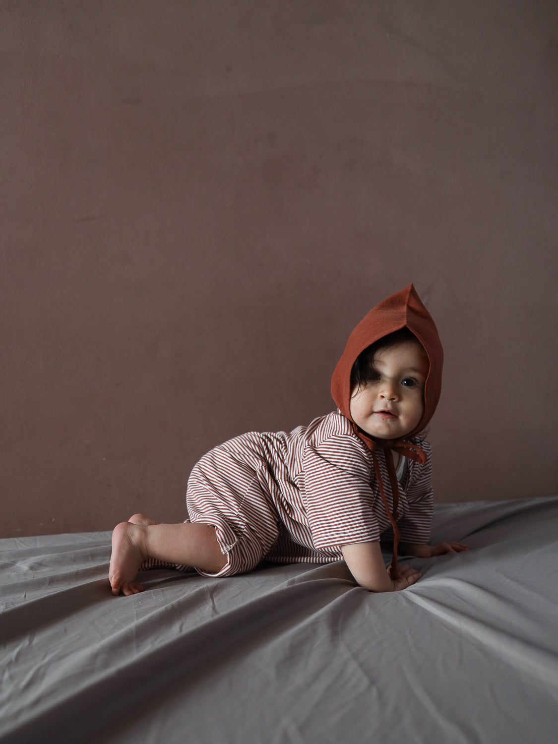Ein Baby trägt den Wickel Overall Baumwolle - Ringel Ziegel, bestehend aus einem rot-weiß gestreiften Einteiler, liegend auf einer grauen Decke, mit neugierigem Blick in die Kamera.