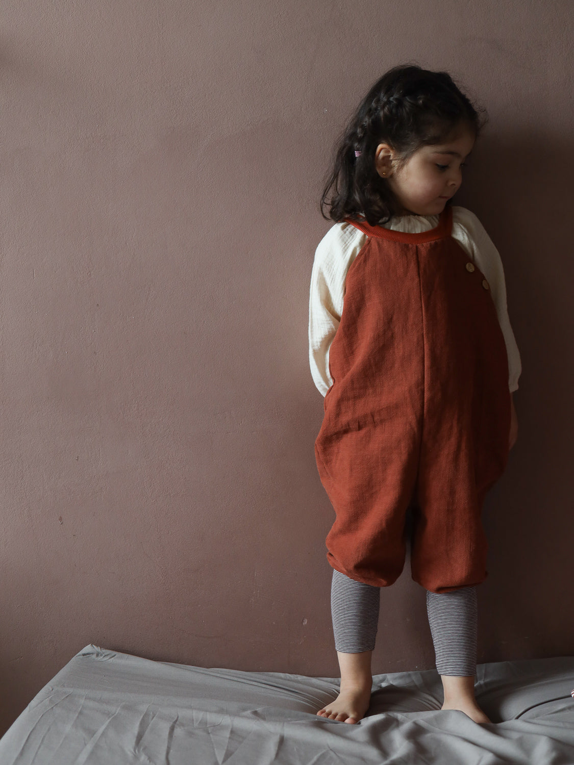 Kleinkind trägt die Schlupflatzhose Leinen - Ziegel, kombiniert mit einem cremefarbenen Langarmshirt und gestreiften Leggings, stehend auf einem Bett. Die Latzhose zeigt einen lockeren, komfortablen Schnitt.
